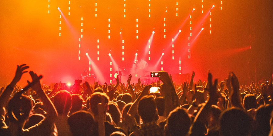 group of people in front of a stage with orange lighting, others holding their gadgets while others raising their hands in the air