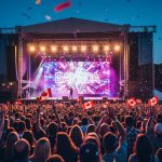Outdoor summer festival at night with a glowing main stage, colorful lights, raised hands, and confetti over a Canadian park setting.